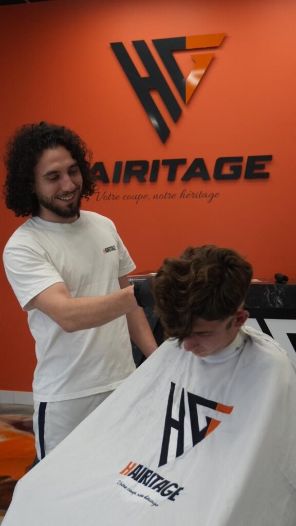 Coiffeur Hairitage souriant devant le mur orange au logo